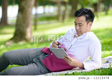 Young man in causal using mobile device tablet iPad laying on a mat in a park 11213478