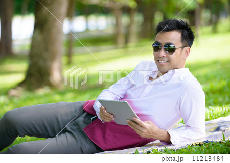 Young man in causal using mobile device tablet iPad laying on a mat in a park 11213484