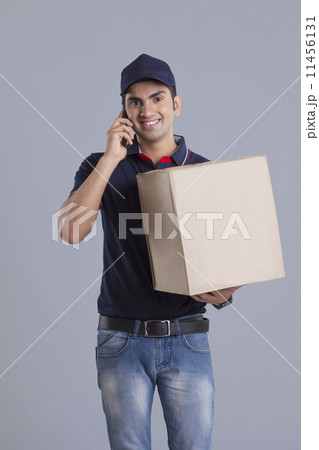 Portrait of delivery man carrying package while answering mobile phone against gray background 11456131