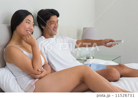 Smiling asian couple lying on bed watching tv 11526731