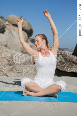 Blonde woman sitting in lotus pose on beach on mat with arms rai 11531778
