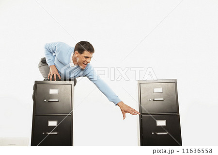 Businessman on top of filing cabinet reaching for other filing cabinet Businessman on top of filing cabinet reaching for other filing cabinet 11636558