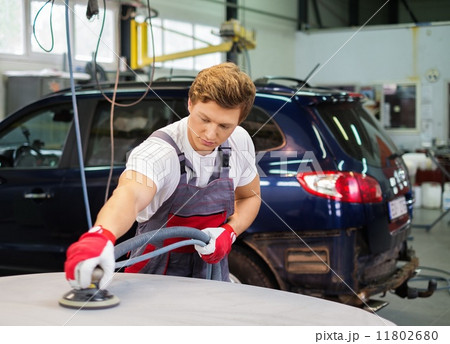 Young serviceman performing grinding with machine on a car bonnet in a workshop 11802680