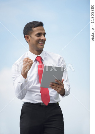 Successful young Indian corporate executive victory using iPad mobile device tablet along promenade Successful young Indian corporate executive victory using iPad mobile device tablet along promenade 11805880