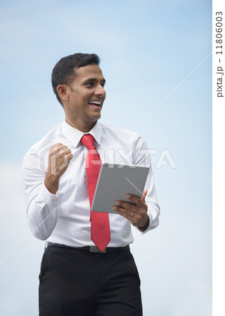 Successful young Indian corporate executive victory using iPad mobile device tablet along promenade Successful young Indian corporate executive victory using iPad mobile device tablet along promenade 11806003
