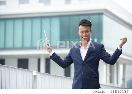 Successful young chinese businessman victory gesture using iPad tablet front of glass building Successful young chinese businessman victory gesture using iPad tablet front of glass building 11810157