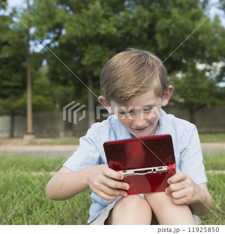 A young boy sitting on the grass playing a hand held electronic game.  11925850