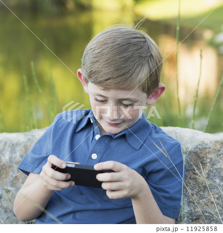 A young boy outdoors sitting leaning against a rock, using a handheld electronic game. A young boy outdoors sitting leaning against a rock, using a handheld electronic game. 11925858