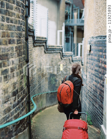 A woman walling down a narrow street, pulling a suitcase and holdin an orange backpack. 11925893