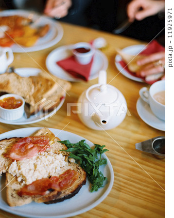 A table covered with plates of food. A cooked breakfast of bacon, eggs and toast and pot of tea. Two people seated 11925971