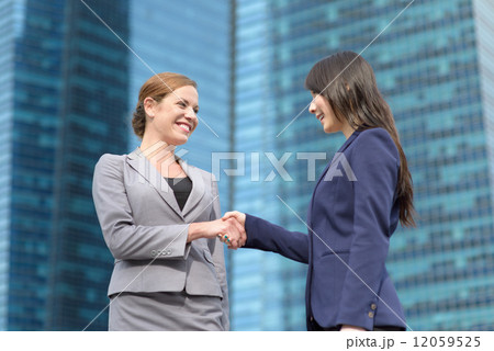 Two young successful business women shaking hands in front of blue glass office building 12059525