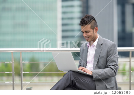 One successful young Malay business man using laptop along promenade in front of office building  12065599