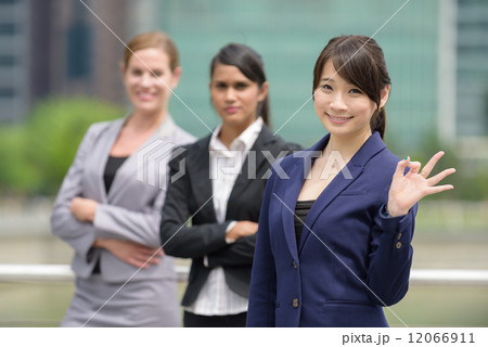 Team leader of 3 young successful mixed race business women giving okay sign in front of office 12066911