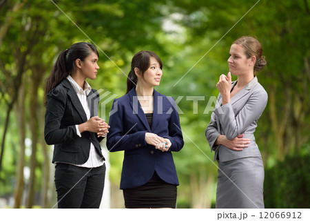 Three young successful mixed race business women chatting along green walkway 12066912