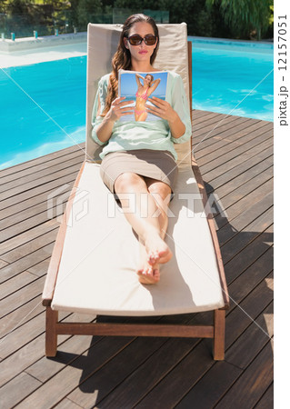 Woman reading book on sun lounger by pool 12157051