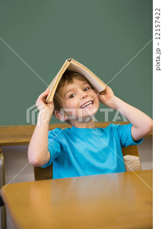 Happy pupil holding book on his head at desk 12157422