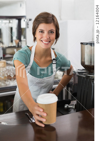 Pretty barista smiling at camera holding disposable cup Pretty barista smiling at camera holding disposable cup 12164004