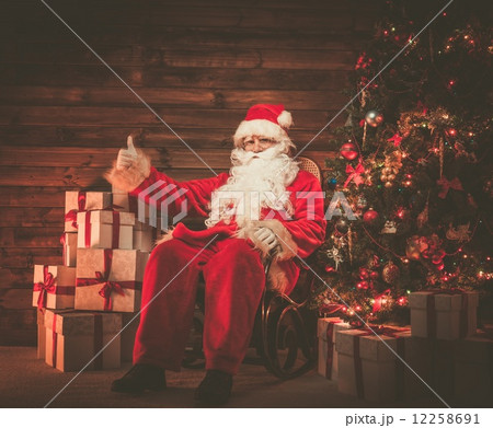 Santa Claus sitting on rocking chair in wooden home interior with gift boxes around him Santa Claus sitting on rocking chair in wooden home interior with gift boxes around him 12258691