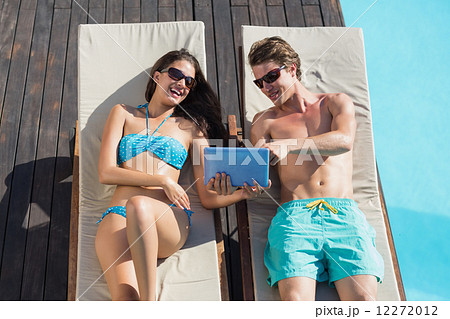 Couple using digital tablet on sun loungers by swimming pool 12272012