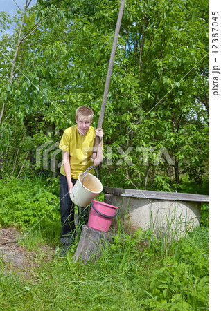 The boy, the teenager, pours water in a bucket from a well 12387045