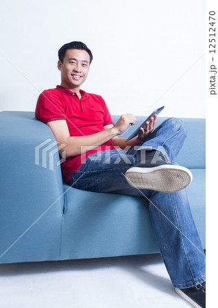 A young Asian man enjoy the moment using his mobile device while seating in his living room A young Asian man enjoy the moment using his mobile device while seating in his living room 12512470