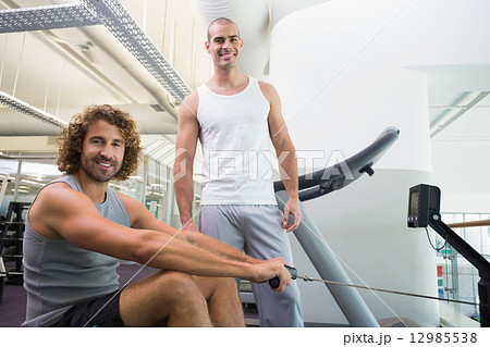 Male trainer assisting man on fitness machine at gym Male trainer assisting man on fitness machine at gym 12985538