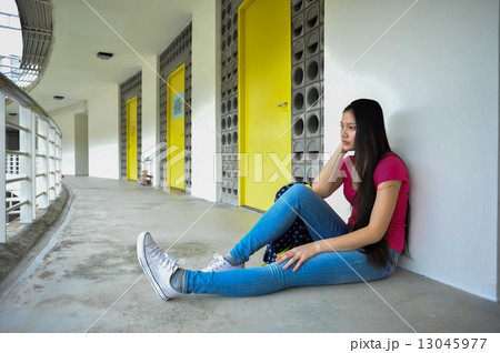 A sad and lonely young Asian female student seating by herself at a corridor of a campus A sad and lonely young Asian female student seating by herself at a corridor of a campus 13045977