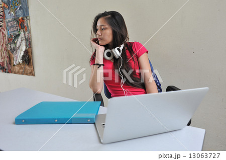 A young and beautiful Asian female student pondering / brainstorming at her desk in front of laptop A young and beautiful Asian female student pondering / brainstorming at her desk in front of laptop 13063727