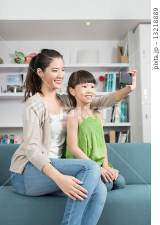 Young chinese mother and daughter taking selfies using mobile device smart phone relaxing on a sofa 13218889