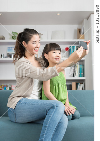 Young chinese mother and daughter taking selfies using mobile device smart phone relaxing on a sofa 13218890
