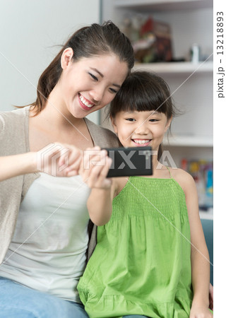 Young chinese mother and daughter taking selfies using mobile device smart phone in the living room 13218893