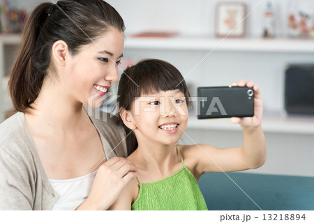Young chinese mother and daughter taking selfies using mobile device smart phone in the living room 13218894