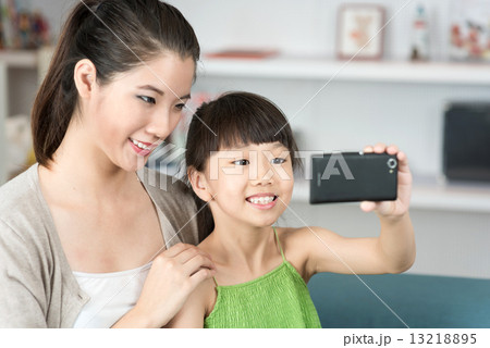 Young chinese mother and daughter taking selfies using mobile device smart phone in the living room 13218895