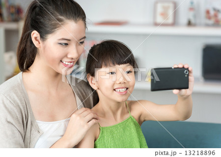 Young chinese mother and daughter taking selfies using mobile device smart phone in the living room 13218896