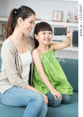 Young chinese mother and daughter taking selfies using mobile device smart phone relaxing on a sofa 13218897