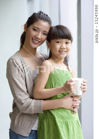 Young chinese mother and daughter holding a cup of hot chocolate at hand along glass window 13245998