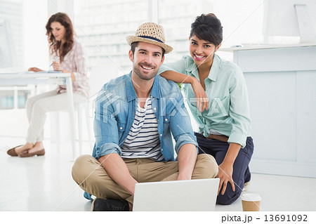 Smiling colleagues sitting on the floor using laptop 13691092