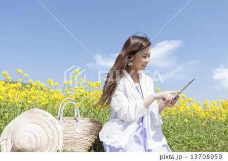 菜の花畑に座ってタブレットPCを操作している笑顔の女性 13708959