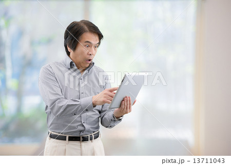 自宅リビングの窓際でタブレットを操作しているカジュアルウェアー姿の中年男性 13711043