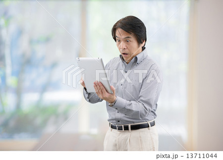 自宅リビングの窓際でタブレットを操作しているカジュアルウェアー姿の中年男性 13711044
