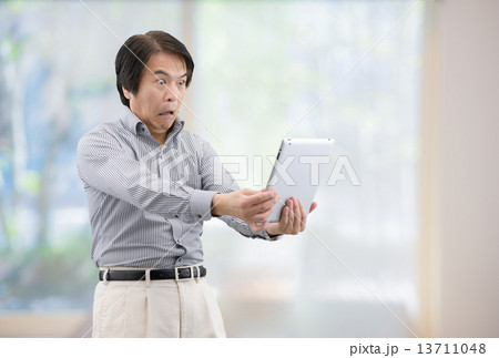 自宅リビングの窓際でタブレットを操作しているカジュアルウェアー姿の中年男性 13711048