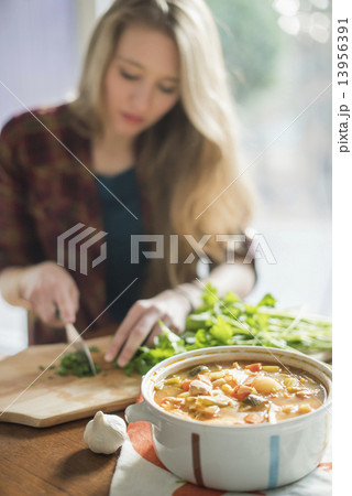 A woman sitting at a table, chopping herbs, a bowl of vegetable stew in the foreground. 13956391