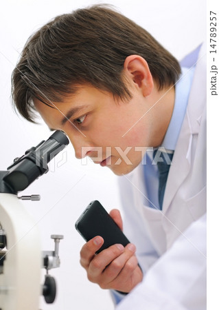 Researcher looking in microscope and making notes on voice recor Researcher looking in microscope and making notes on voice recor 14789257