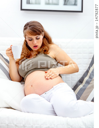 Surprised beautiful pregnant woman sitting on sofa and holding cup of tea in hand. 14792877