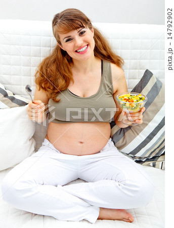 Smiling beautiful pregnant female sitting on sofa with fruit salad in hand. 14792902