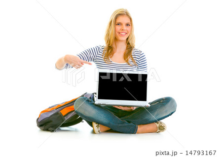 Smiling teengirl sitting on floor with schoolbag and pointing finger on laptop with blank screen. 14793167