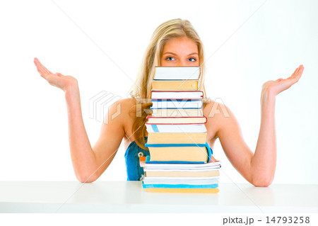 Sitting at table confused teen girl spreading hands apart and looking out of pile of book. 14793258