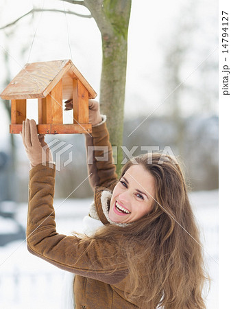 Smiling young woman hanging bird feeder on tree in winter outdoo 14794127