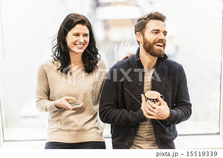 A business woman seated by a window holding a smart phone, talking to a man holding a coffee cup. 15519339