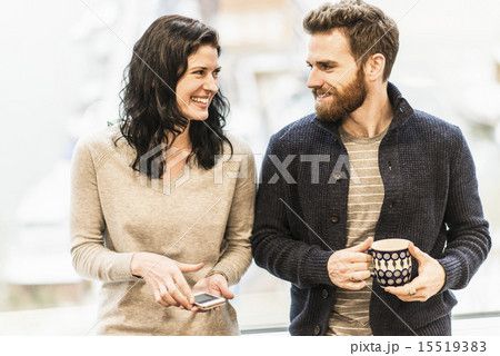 A business woman seated by a window holding a smart phone, talking to a man with a coffee cup. A business woman seated by a window holding a smart phone, talking to a man with a coffee cup. 15519383